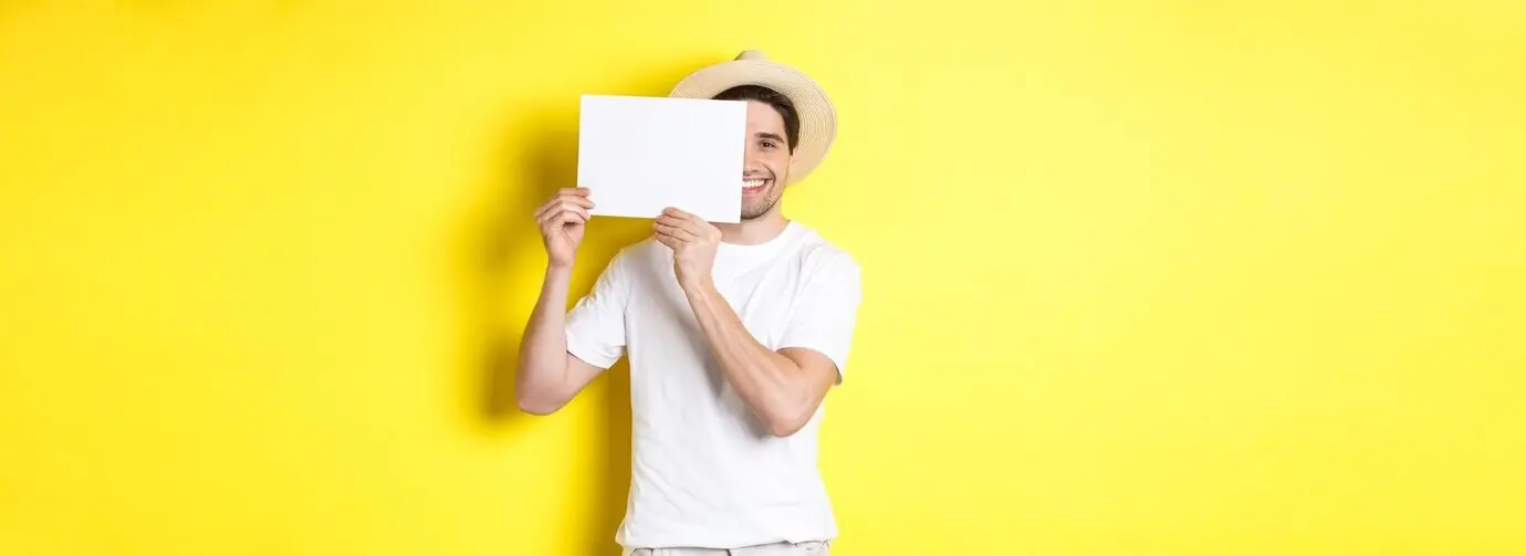 Счастливый парень в отпуске показывает пустой лист бумаги под ваш логотип, держит его у лица и улыбается.