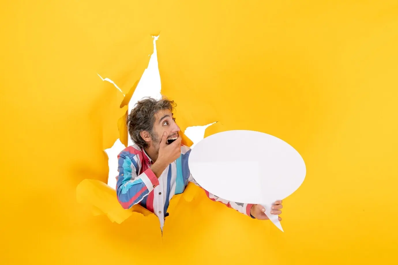 Вид сверху на удивлённого молодого мужчину, указывающего на белый лист с пустым пространством в рваном отверстии в жёлтой бумаге.